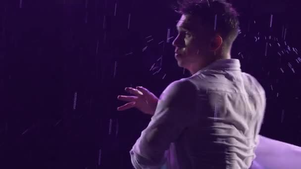 Emotional performance of contemporary ballet choreography in the rain in dark studio with blue light. Silhouette of wet man gracefully moving and performing acrobatic elements. Slow motion. Close up. - Video, Çekim