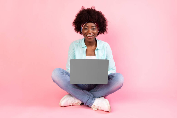 ピンクの背景の上に座ってノートパソコンを使用して幸せなアフリカ系アメリカ人女性 - 写真・画像