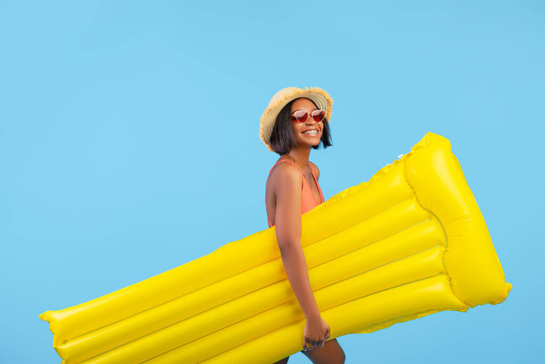 Young black lady in swimwear and straw hat holding yellow inflatable lilo on blue studio background - Photo, Image