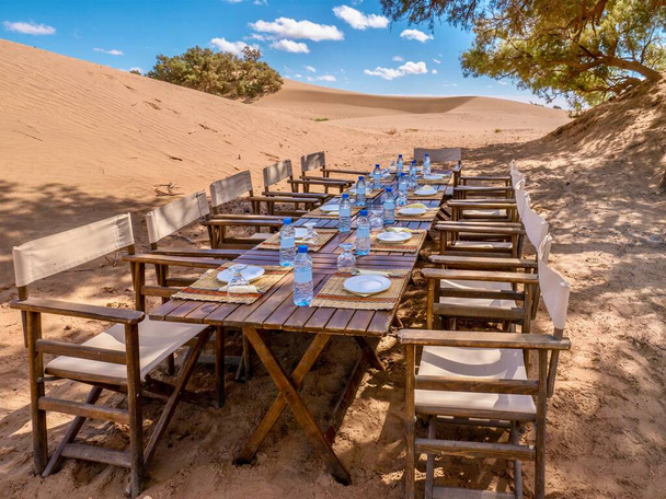 A long wooden table and chairs prepared for a tourist lunch in the Sahara Desert, Morocco. Conceptual for last minute tourist and travel cancellations or the environmental issue of plastic water bottles. - Photo, Image