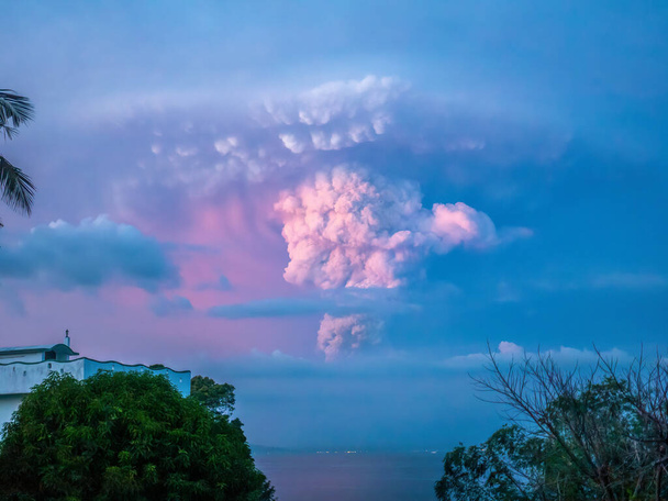 Вулкан Таал, Филиппины - 12 января 2020 года. Вулкан Таал извергается во время заката, его шлейф виден в небе над огнями города Батангас, вид с острова Миндоро, в 33 милях от извержения. - Фото, изображение