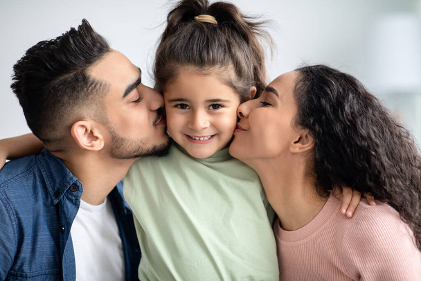Glückliche Kindheit. Liebevolle arabische Eltern küssen ihre kleine Tochter auf beide Wangen - Foto, Bild