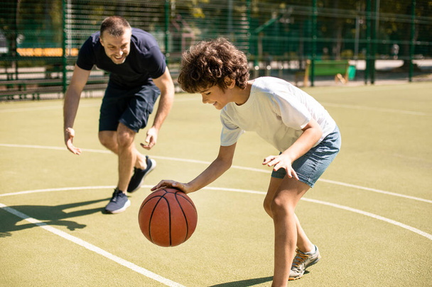 Ο αθλητικός μπαμπάς μαθαίνει στο γιο του να παίζει μπάσκετ έξω. - Φωτογραφία, εικόνα