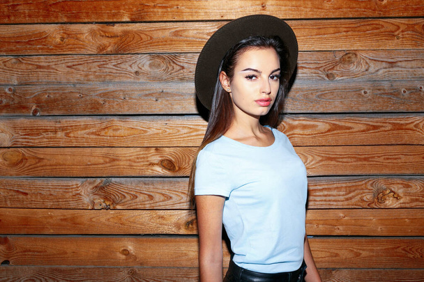 Retrato de cerca de una chica elegante con sombrero, su cabello recto vestido con camiseta azul cerca de la pared de madera, mirando a la cámara. - Foto, Imagen
