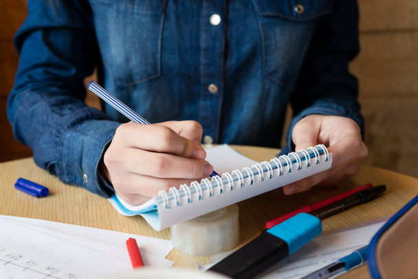 Girl in a blue denim shirt sitting in a cafe makes notes in a notepad with a pen. A sheets with various charts, adhesive tape - Photo, image