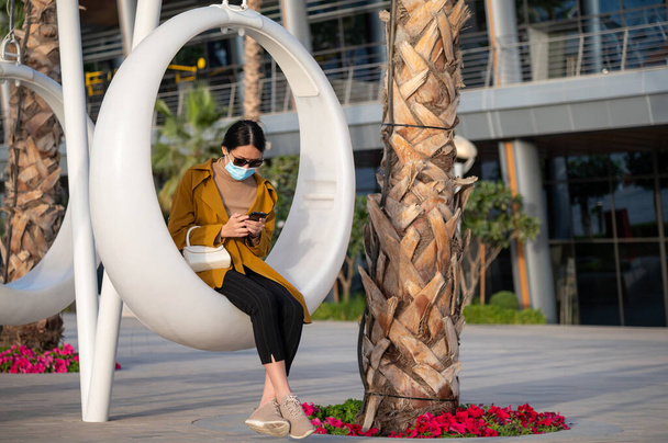 Yalnız bir Asyalı kadın açık havada salıncakta telefon kullanıyor maske takıyor ve toplum içinde mesafeli davranıyor. - Fotoğraf, Görsel