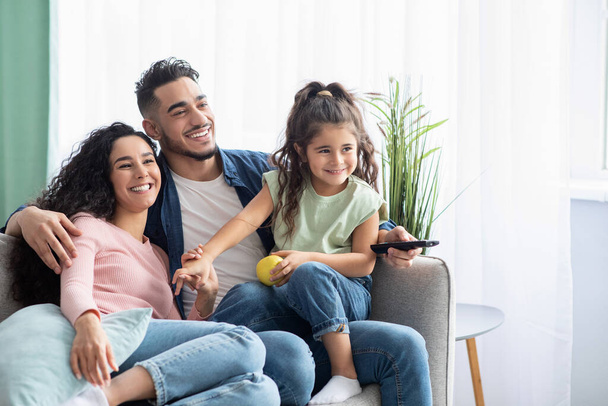 Familieweekend. Vrolijke Arabische ouders en kleine dochter thuis tv kijken - Foto, afbeelding