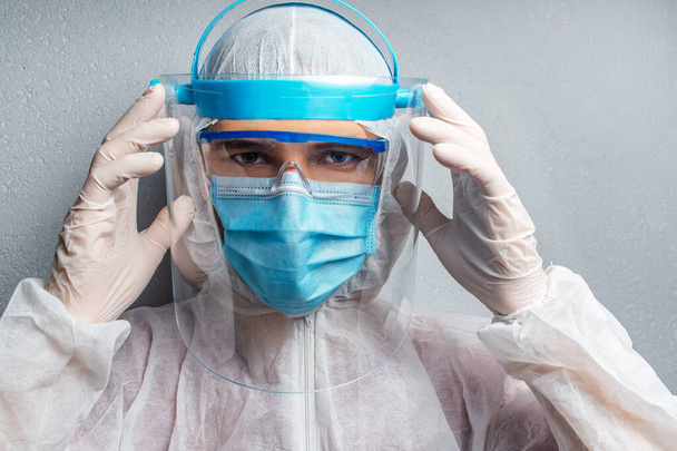 Studio portrait of young doctor man holding face shield with hands, wearing PPE suit against coronavirus and covid-19, on background of textured wall. - Photo, image