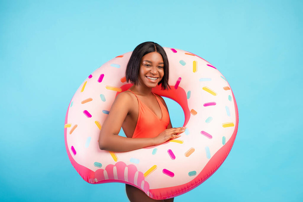Portrait of happy millennial black lady in swimwear standing with donut shaped inflatable ring on blue studio background - Photo, image