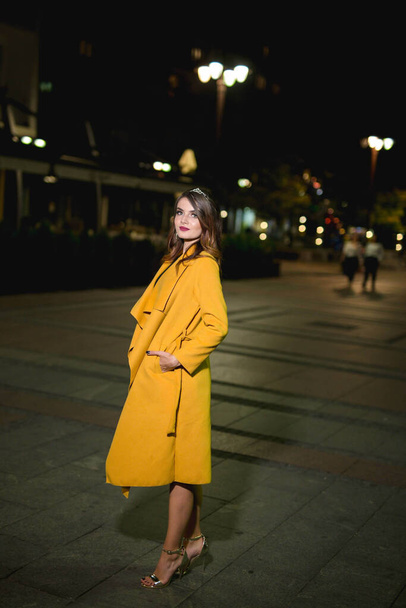 A beautiful and elegant Caucasian woman in a dress and a yellow coat posing on a street at night - Zdjęcie, obraz