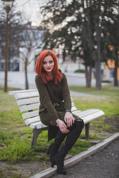 An attractive young Caucasian female with red hair sitting on a bench in a park - Foto, afbeelding