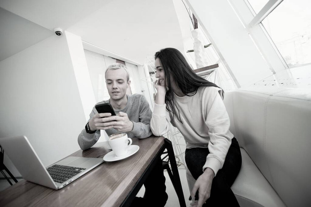 Young couple meets in coffeehouse. Blonde man showing something fun in his smartphone to brunette attractive woman. Modern laptop on table. Social media concept. Woman propped on her hand and smiles - Photo, Image