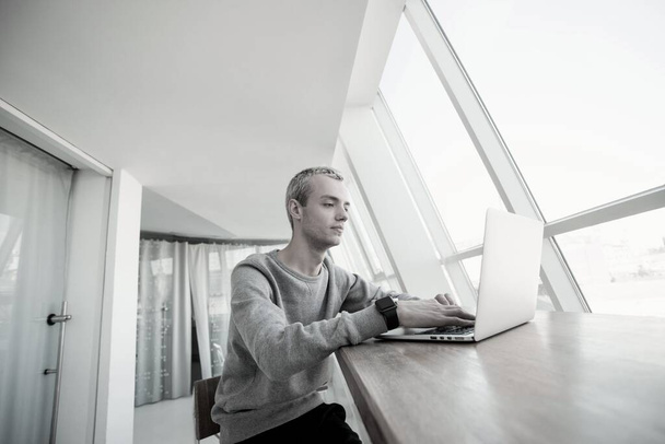 Geconcentreerde freelancer Geconcentreerde freelancer met laptop werkt voor een helder panoramisch venster. Goed uitziende jongeman in casual grijze sweatshot kijkt in het scherm van zijn computer. Samenwerking - Foto, afbeelding