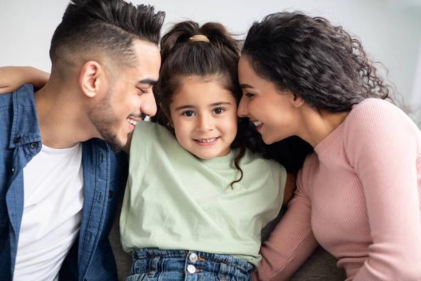 Rodičovská láska. Šťastní Arabští rodiče mazlící se se svou roztomilou dceruškou doma - Fotografie, Obrázek
