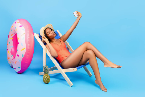 Sexy black woman sitting in lounge chair, taking selfie with smartphone over blue studio background, full length - Photo, Image