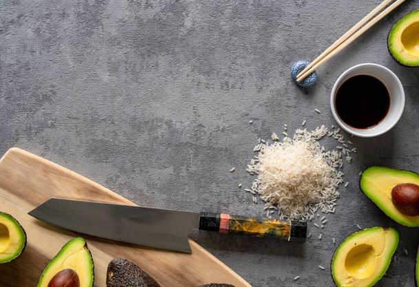 A top view of fresh avocados, a cutting board and knife, rice, and chopsticks on a grey surface, copy space - Fotografie, Obrázek