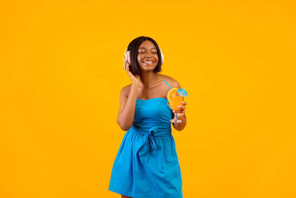 Summer party concept. Sexy black woman with tasty cocktail listening to music in headphones on orange studio background - Φωτογραφία, εικόνα