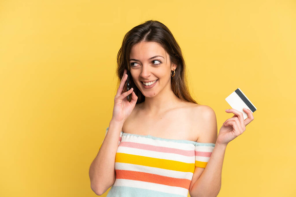 黄色の背景に隔離された若い白人女性携帯電話との会話を維持し、クレジットカードを保持 - 写真・画像
