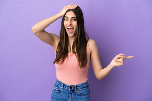 Young caucasian woman isolated on purple background surprised and pointing finger to the side - Foto, afbeelding