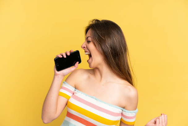 Young caucasian woman isolated on yellow background using mobile phone and singing - Photo, image