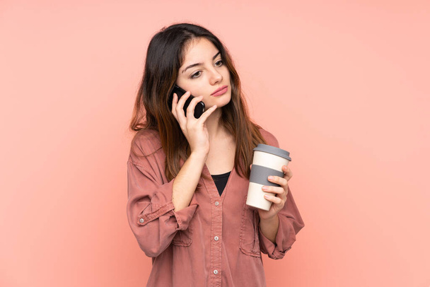 Junge kaukasische Frau isoliert auf rosa Hintergrund hält Kaffee zum Mitnehmen und ein Handy - Foto, Bild