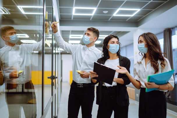 Business colleagues  in protective face mask writing on sticky note on glass wall with coworkers standing by in office. Startup Business. Planning, analysis. Covid-2019. - Foto, Imagem