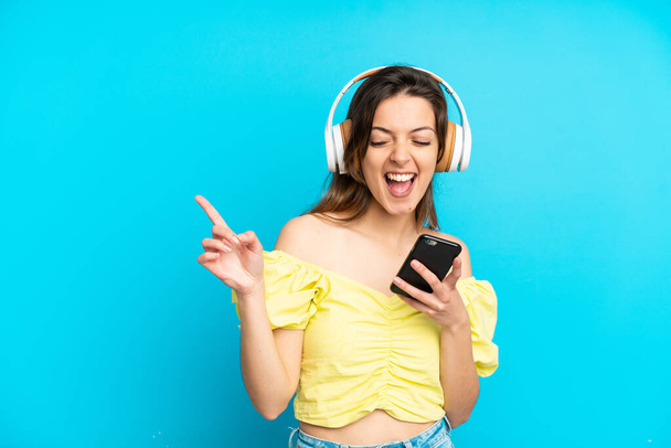 Genç beyaz kadın mavi arka planda izole edilmiş. Cep telefonuyla müzik dinliyor ve şarkı söylüyor. - Fotoğraf, Görsel