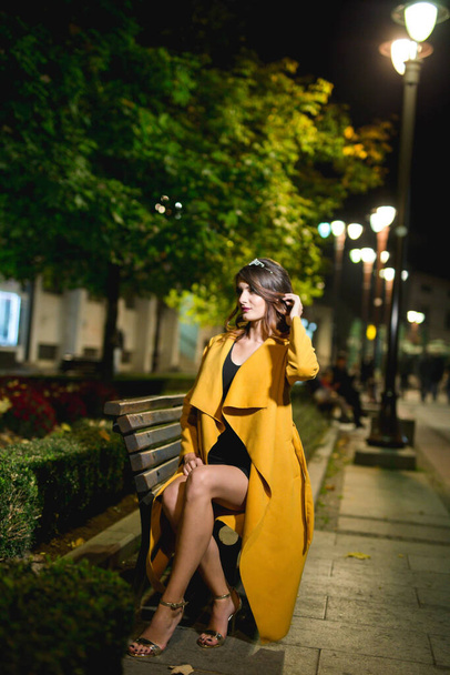 An attractive Caucasian female in a yellow coat sitting on a bench, posing in a park in the evening - Foto, afbeelding