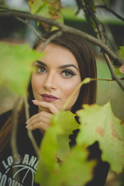 A young brunette Caucasian female posing near a tree with green leaves in a park - 写真・画像