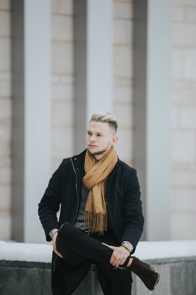A handsome man in a black winter outfit with a brown scarf sitting on a stone in front of the building - Fotoğraf, Görsel