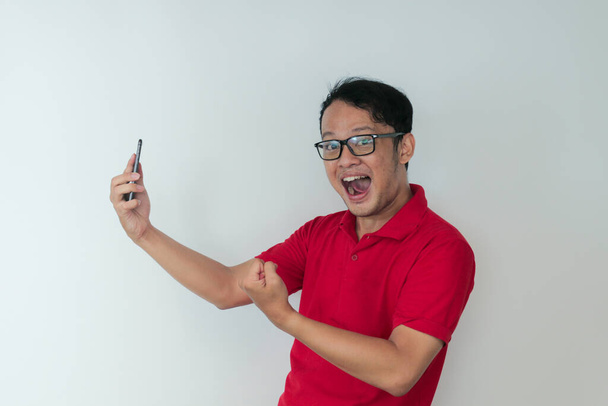 Young Asian man shocked and happy with what he see in the smartphone on isolated grey background. - Valokuva, kuva