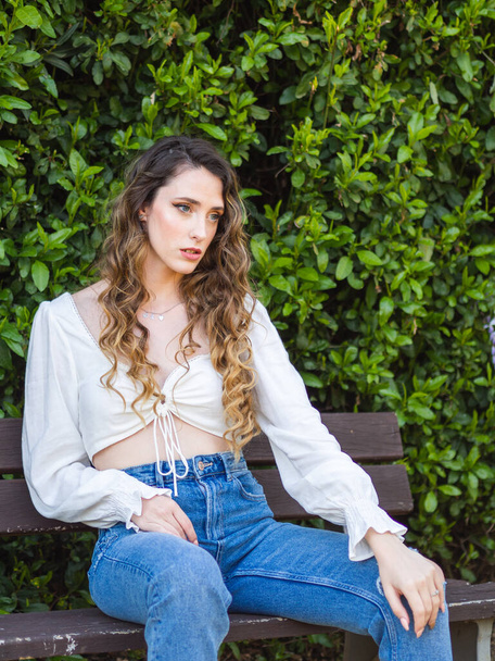 A young Spanish female wearing jeans and a white blouse is sitting on a park bench; springtime - Foto, Imagen