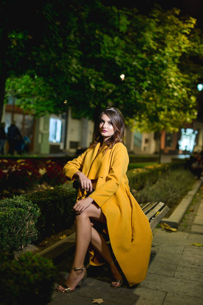 A beautiful and elegant Caucasian woman in a black dress and a yellow coat posing on a street at night - Fotografie, Obrázek