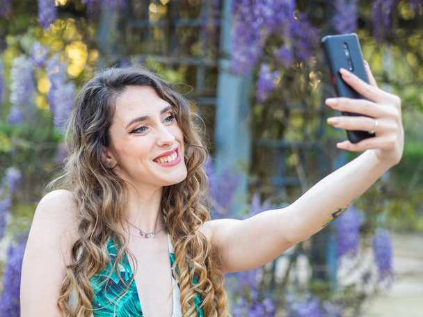 A happy Spanish female with long curly hair is taking a selfie in the blooming spring park - Φωτογραφία, εικόνα