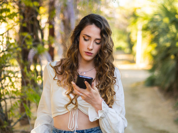 A young Spanish female is using a smartphone while walking in a spring park - Foto, imagen