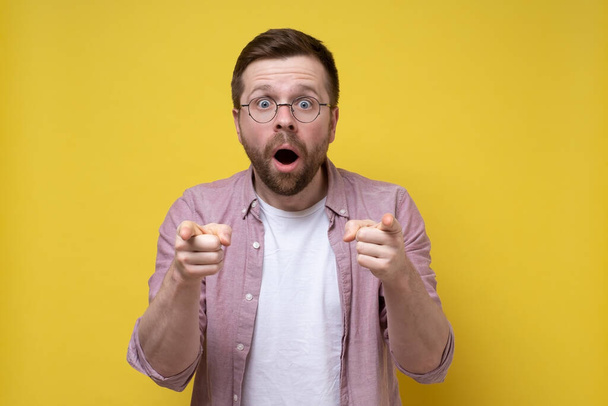 Surprised man with glasses points with index fingers and looks with large eyes, with an open mouth. Yellow background. - Photo, Image