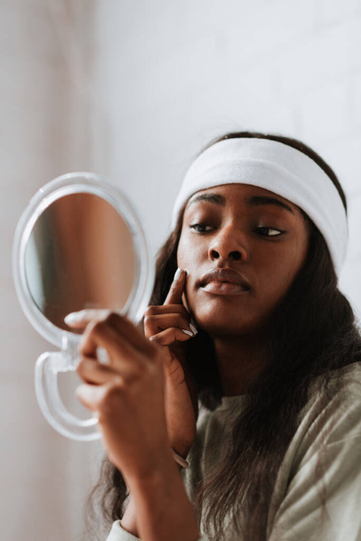 A beautiful young African female wearing a white headband looking at herself in a hand mirror in her bedroom - Foto, Bild