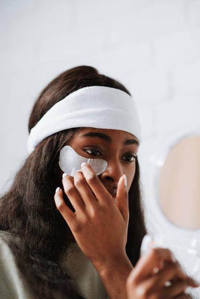 A young African female applying under-eye recovery patches at home while looking in a hand mirror - Фото, изображение