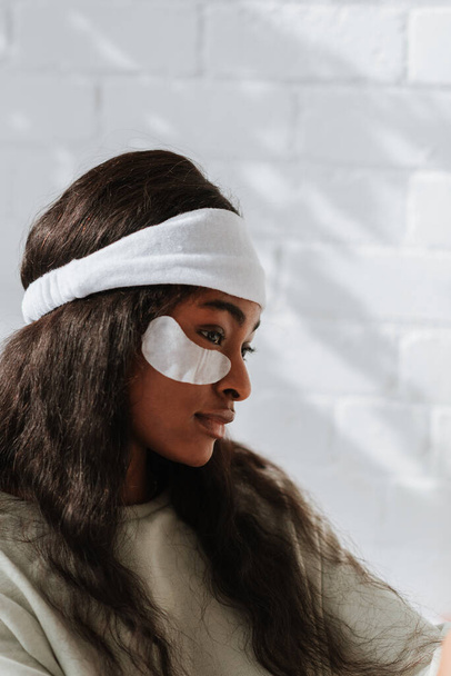 A side view of a young African female wearing a white headband with under-eye recovery patches at home - Foto, imagen