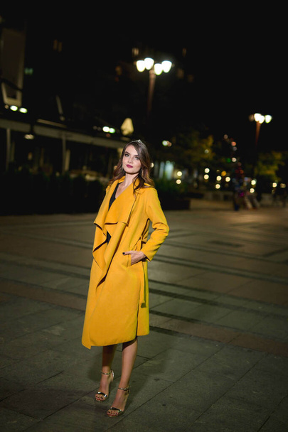 A beautiful and elegant Caucasian woman in a black dress and a yellow coat posing on a street at night - Foto, imagen
