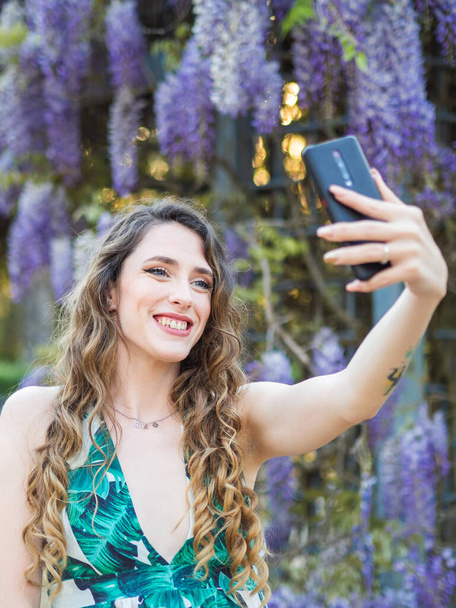 A beautiful happy woman taking a selfie in the background of violet blooming flowers - Foto, imagen
