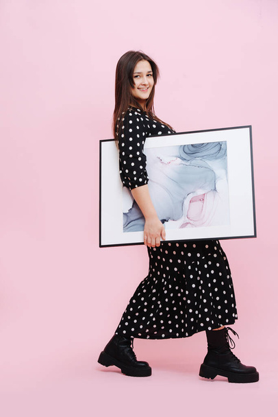 Funny social young woman in polka dot dress carrying big modern abstract painting under her hand. She's wearing heavy boots, tilting back while walking. Looking at the camera. Over pink background. - Fotografie, Obrázek