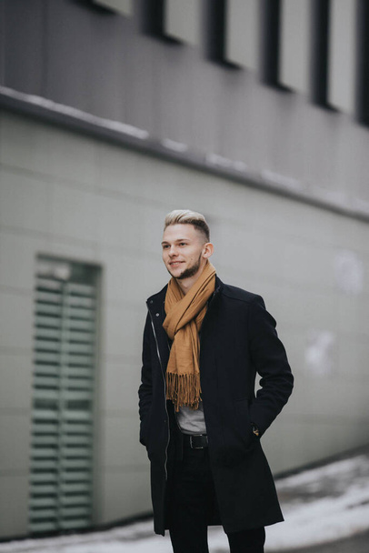 A handsome young smiling man in a black winter outfit withtrendy brown scarf standing on a wet street - Foto, Imagem
