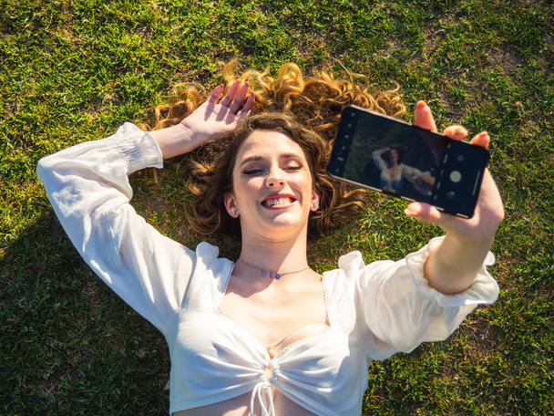 A top view of a young Spanish girl taking a selfie while relaxing on the grass in the spring park - Φωτογραφία, εικόνα