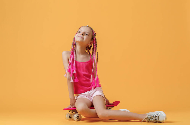 Active young girl in pink shirt and shorts sitting on skateboard - 写真・画像