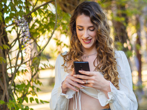 A long-haired young Spanish lady is using a smartphone while posing in the spring park - Photo, image