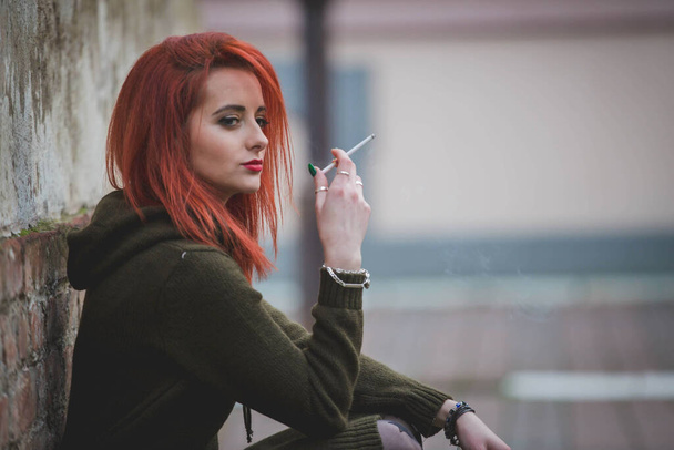 A red-haired young female in a beautiful winter dress smoking in front of a brick wall - Φωτογραφία, εικόνα