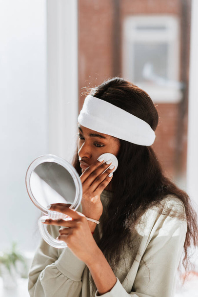 An attractive young African female cleaning her face with a cotton disc before going to bed looking in a hand mirror - Фото, изображение