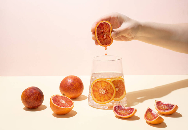 Samičí ruka vtlačuje polovinu zralého šťavnatého pomeranče do sklenice funkční vody s citrusovými plátky na žlutorůžovém pozadí v jasném světle. Čerstvý džus kape dolů. - Fotografie, Obrázek
