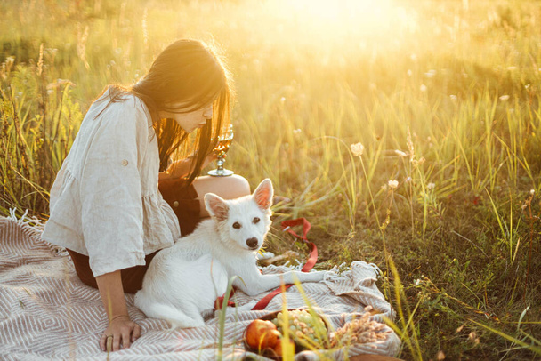 夏の牧草地で暖かい日当たりの良い光の中で毛布の上で白い犬とリラックスしたスタイリッシュな女性。休暇とペットとピクニック。若いですboho女性楽しんで夕日とともにスイスの羊飼い子犬 - 写真・画像